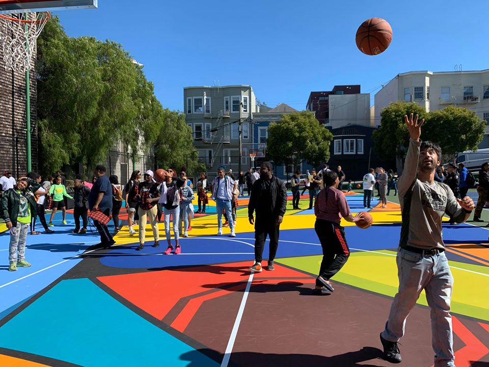 People mingle on newly painted basketball court mural. One person is throwing a basketball in the air.