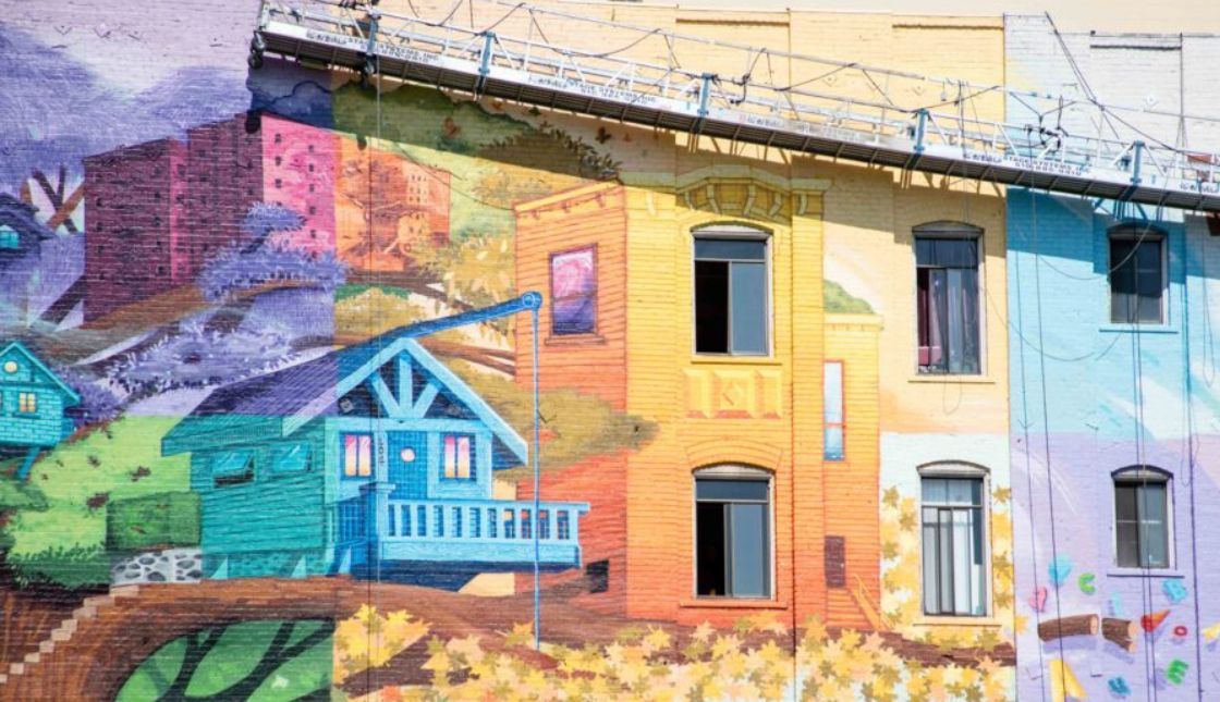 An image of a mural painted on the side of a large building. The mural depicts a tree house in front of an apartment building in rainbow colors.