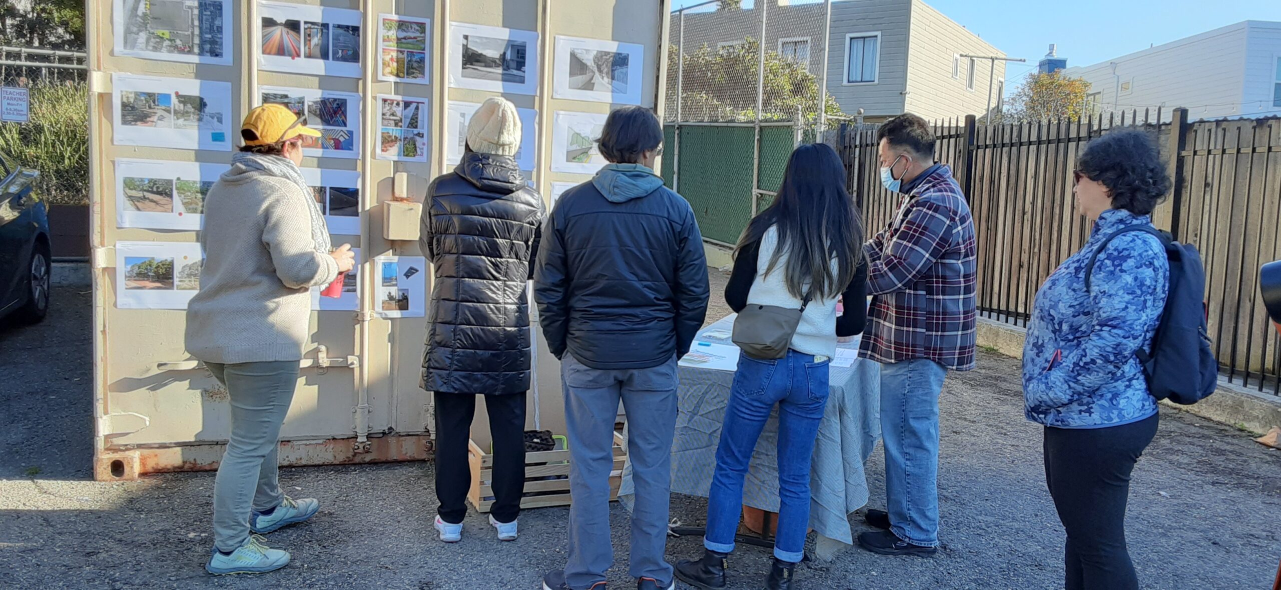Six community members in cold weather clothes review design ideas on sheets of paper affixed to a storage container.